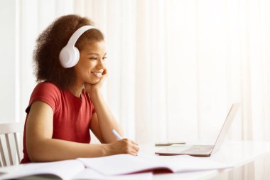 Uzaklık Eğitimi. Gülümseyen siyahi kadın portresi, dizüstü bilgisayar kullanıyor ve deftere yazıyor, not alıyor, özel ders veya webinar izliyor, evde ders çalışıyor, ekranlara bakıyor.