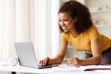 Gülümseyen Afro-Amerikan kadın masasının üzerine eğiliyor, dizüstü bilgisayara yazıyor ve notlar alıyor, neşeli siyah kadın odaklanıyor ve evde öğrenim ortamı ile meşgul.