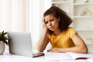 Sıkıcı iş. Ofiste dizüstü bilgisayarda çalışan siyahi bir kadın üzgün bir Afrikalı Amerikalı kadın bilgisayarla masada oturuyor ve canı sıkkın bir yüz ifadesiyle ekrana bakıyor.