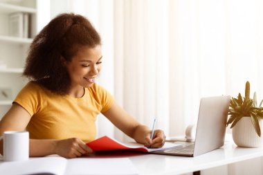 Gülümseyen siyah kadın dizüstü bilgisayarla çalışıyor ve not defterine not alıyor, masada oturan ve bilgisayardan bilgi alan mutlu, genç Afrikalı Amerikalı kadın,