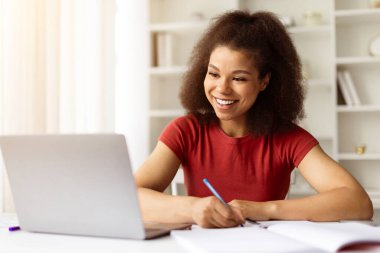 Sırıtkan Afrikalı genç bir kadın dizüstü bilgisayarda çalışıyor ve not alıyor, mutlu siyah kadın online çalışıyor, web sitelerini izliyor, bilgisayar kullanıyor ve deftere yazı yazıyor, yakın çekim yapıyor.