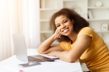 Neşeli Afrikalı Amerikalı kız öğrenci dizüstü bilgisayarla masaya yaslanıyor ve kameraya gülümsüyor, pozitifti güzel siyahi kadın okuldan sonra dinleniyor ya da çalışıyor, uzaktan işten ya da eğitimden keyif alıyor, boşluğu kopyalıyor.