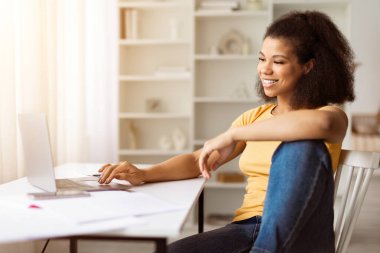 Neşeli genç siyah kadın dizüstü bilgisayar kullanıyor ve evdeki rahat poz veriyor, mutlu Afrikalı Amerikalı kadın internette takılıyor, internette sörf yapıyor, bilgisayarla evde dinleniyor.