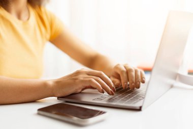 Ofis ortamında dizüstü bilgisayarda yazan genç Afrikalı Amerikalı kadınların elleri, parmakları klavyede dinlenirken, akıllı telefon da okumak veya uzaktan çalışmak için ideal beyaz masanın üzerinde yatıyor.