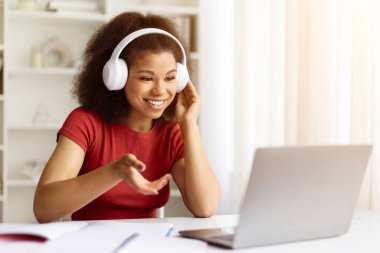 Video görüşmesi için dizüstü bilgisayar kullanan, kablosuz kulaklık takan mutlu genç siyahi kadın, Afrikalı Amerikalı kız öğrencilerin internet dersi ya da giysisi var, evdeki masasında oturuyor, uzay kopyası yapıyor.