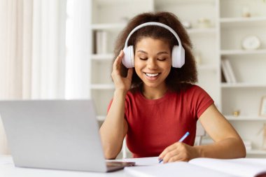Neşeli siyah kadın kulaklıkla müzik dinliyor ve evde masa başında otururken not alıyor, mutlu Afrikalı Amerikalı kadın öğrenci dizüstü bilgisayarla çalışıyor, çevrimiçi eğitimin tadını çıkarıyor.