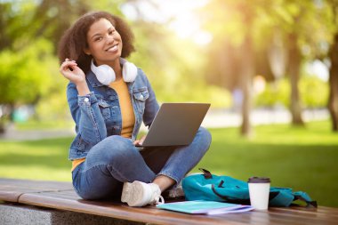 Dışarıda kucağında dizüstü bilgisayarla oturan mutlu siyahi bir kadın, kulaklık takan ve kampüste kot ceket giyen neşeli bir Afro-Amerikan kadın, kameraya gülümsüyor.