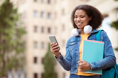 Smiling African American female student with smartphone in hand walking on campus, happy black young woman holding notebooks and checking messages on cellphone, enjoying modern communication