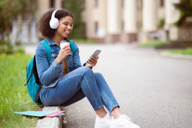 Zeki telefon kullanan ve dışarıda kahve içen mutlu siyah öğrenci kampüste dinlenen kablosuz kulaklık takan neşeli Afrika kökenli Amerikalı kadın.