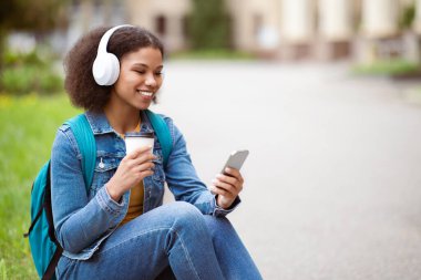 Öğrenci eğlencesi. Genç siyah kadın akıllı telefon kullanıyor ve açık havada kahve içiyor, mutlu Afro-Amerikan kadın cep telefonunda tarama uygulaması yapıyor ya da video görüşmesi yapıyor, uzay boşluğunu kopyalıyor.