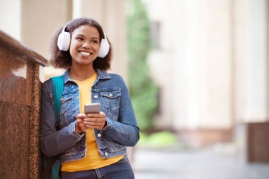 Kampüste dinlenirken Smartphone 'da müzik dinleyen gülümseyen zenci bir kadının portresi, Kablosuz kulaklık takan pozitif Afrikalı genç Amerikalı kadın,