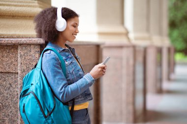 Genç Afrikalı Amerikalı kız öğrenci dışarıda duruyor, gülümsüyor ve akıllı telefonu kontrol ediyor, bina direğine yaslanıyor, beyaz kulaklıklardan müzik dinliyor, kampüs sahnesinde dinleniyor.