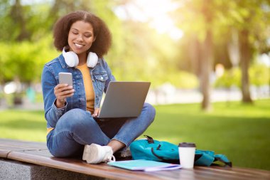 Neşeli Afro-Amerikalı kız öğrenci açık havada birden fazla iş yapıyor, akıllı telefon ve laptop kullanıyor, mutlu siyah kadın kampüs ortamında bankta bağdaş kurarak oturuyor.