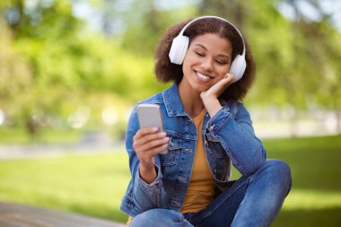 Afro-Amerikalı genç bayan öğrenci dışarıda, kulaklık takıyor ve sıcak bir şekilde gülümsüyor. Güneşli kampüs ortamında akıllı telefona bakıyor, müzik dinliyor ya da internette geziniyor.