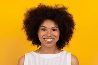 Gülümseyen, kıvırcık saçlı, beyaz kolsuz bluz giyen, parlak sarı stüdyo arka planında duran mutlu siyah kadın.