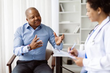 Siyahi erkek hasta, klinikteki toplantı sırasında Doktor Kadın 'la konuşuyor, Afrikalı Amerikalı olgun bir adam terapist kadına sağlık sorunlarını anlatıyor ve panoya kayıt yapıyor.
