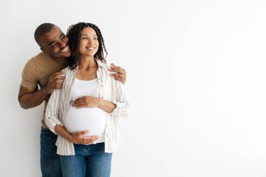 Siyah hamile çift fotokopi uzayına sarılıp yan bakıyor, Afrikalı genç koca ve karısı beyaz arka planda bebek pozu bekliyor.