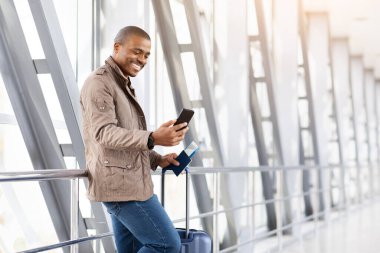 Genç siyahi adam havaalanı terminalinde telefonu kontrol ederken gülümsüyor, yeni uygulamalara bakıyor, günlük kıyafetler giyiyor, doğal ışık ve dijital yaşam tarzıyla samimi seyahat anları yaşıyor.