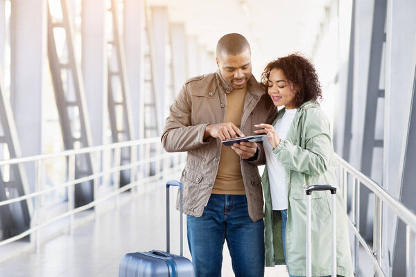 Молодая черная пара рассматривает вместе билеты в аэропорту, имея при себе паспорт и документы, чемодан поблизости, сфокусированный момент подготовки к путешествию или чекину, копировальное пространство