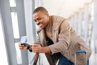 Parlak havaalanı koridorunda seyahat belgelerini incelerken gülümseyen siyahi genç adam, mutlu Afrika kökenli Amerikalı erkek bavulla duruyor, tırabzanlara yaslanıyor ve pasaporta bakıyor, yakın plan