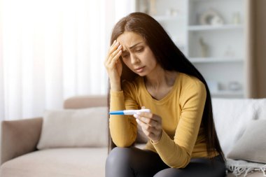 Endişeli genç bir kadın koltukta oturuyor, elinde pozitif hamilelik testi, alnında ellerle, beklenmedik bir şekilde ev ortamını inceliyor, sarı bluzunu giyiyor, boşluğu kopyalıyor.