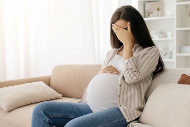 Evde ağlayan üzgün hamile kadın, hamilelik bunalımı geçiren hamile kadın, hamilelik sırasında hormon değişimleri geçiren, oturma odasında oturan umutsuz kadın, elleriyle yüzünü kapatan, boşluğu kopyalayan kadın.