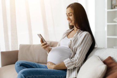 Anneler evdeki kanepeye oturur, bebek karnını nazikçe tutar ve sakin bir gülümsemeyle akıllı telefon kullanır, yumuşak doğal ışıkta sessiz doğum öncesi zamanının tadını çıkarır.