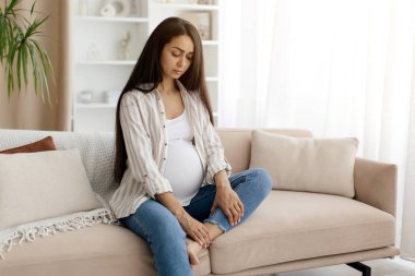 Hamilelik Sağlık Sorunları. İç çamaşırlı hamile bir kadın, şişmiş ayağına masaj yapıyor ve kaşlarını çatıyor. Evdeki kanepede oturan bir kadın bekliyor.