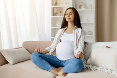 Güzel, hamile bir kadın evde yoga yapıyor oturma odasındaki koltukta nilüfer pozisyonunda oturuyor, müstakbel bir kadın, gözleri kapalı meditasyon yapıyor, sağIık için meditasyon yapıyor.