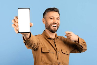 Elinde boş akıllı telefonu işaret eden mutlu genç adam, yakışıklı milenyum erkeği yeni uygulama öneriyor, online teklif sunuyor, mavi arka planda tek başına duruyor,