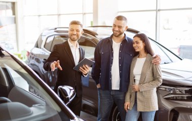 Araba satıcısı galeride genç ve mutlu bir çifte araba sunuyor. Satın alma kararında arkadaş canlısı ve profesyonel atmosfer, aile otomobili seçiyor