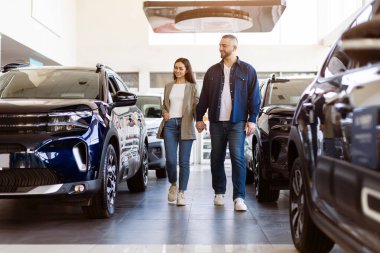 Modern araba galerisinde yürürken el ele tutuşan genç bir çift. Araç alımı sırasında neşeli ve rahat bir atmosfer. Milenyum ailesi otomobili birlikte seçiyor.