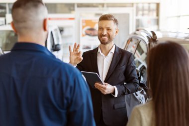 Takım elbiseli, gülümseyen araba satıcısı elinde tablet olan ve seçici bir odak noktası olan modern bir galeride danışmanlık yapan genç bir çifte iyi niyet gösterisi yapıyor.