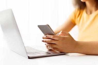 Dizüstü bilgisayarın önünde akıllı telefonu tutan kadınların yakın çekimi. Uzaktan iletişim, çevrimiçi alışveriş veya serbest çalışma veya evde öğrenim sırasında birden fazla iş, kırpıldı