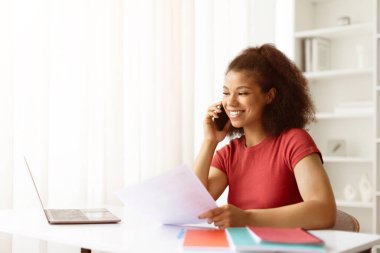 Evden uzaktan çalışan gülümseyen genç siyahi kadın, masadaki belgeleri incelerken telefonla konuşuyor. Pozitif yaşam tarzı, serbest veya uzak çalışma konsepti, kopyalama alanı