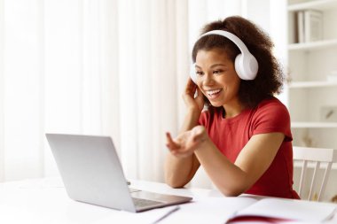 Bilgisayarda uzaktan konuşma sırasında gülümseyen ve el kol hareketi yapan kulaklıklı genç siyahi bir kadın. Konforlu ev ofisinden çevrimiçi öğrenme veya sanal takım iletişimi, boşluğu kopyala