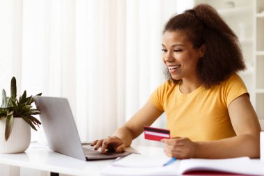 Gülümseyen siyah bir kadın, elinde kredi kartıyla internetten alışveriş yaparken dizüstü bilgisayarda yazı yazıyor. Ev ofisindeki uzak yaşam tarzı ve günlük e-ticaret sahnesi, boşluğu kopyala