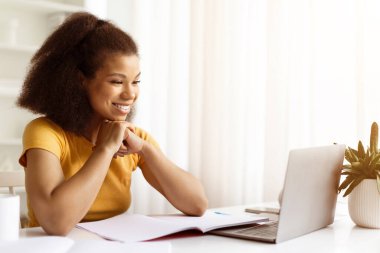 Çevrimiçi derste ya da uzaktan toplantıda dizüstü bilgisayara gülümseyen mutlu siyah kadın. Konforlu ev ortamında çalışan nişanlı ve motive olmuş öğrenci ya da serbest çalışan.