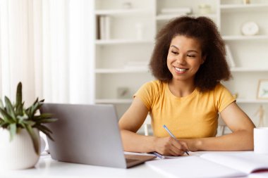 Neşeli genç siyah kadın dizüstü bilgisayara gülümserken not alıyor. Çevrimiçi öğrenime odaklanmış ya da evde bitki dekoru olan rahat bir masada uzaktan çalışmaya odaklanmış, boş alan