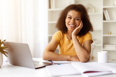 Genç siyahi kadın evde oturuyor, eli yüzü üzerinde, kameraya hafifçe gülümsüyor. Sakin ve dost canlısı uzaktan çalışma ya da rahatlamış iç mekan ortamında çalışma, yakın çekim