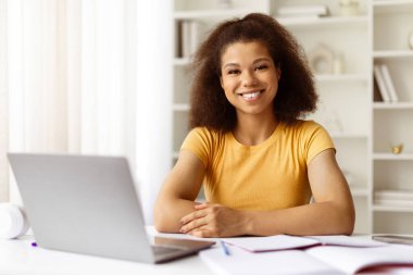 Ev masasında dizüstü bilgisayar ve defterlerle otururken kameraya gülümseyen kendinden emin genç siyahi kadın. Olumlu uzaktan çalışma veya arkadaşça ifade ile öğrenim kavramı, yakın çekim