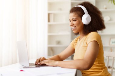 Kulaklık takıp dizüstü bilgisayarda yazan gülümseyen siyahi bir kadın. Müzikten ya da çevrimiçi çalışma sırasında uzaktan iletişimden ya da evden bağımsız çalışmadan hoşlanmak, boşluğu kopyalamak