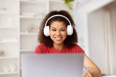 Evde dizüstü bilgisayar kullanan, kulaklıklı, gülümseyen siyah bir kadının ön görüntüsü, sanal sınıfa ya da internet toplantısına katılan mutlu bir kadın, uzaktan çalışma ortamı, yakın çekim.