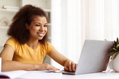 Gülümseyen genç siyahi kadın çevrimiçi ders sırasında ya da uzaktan çalışırken laptop ekranına bakıyor. Konforlu modern ofis kurulumunda çalışma ya da çalışma rutini, kopyalama alanı