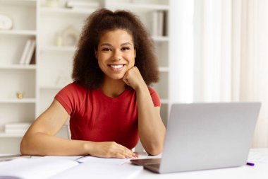 Genç siyahi kadın evinde dizüstü bilgisayarla masasında güven içinde oturuyor. Sakin, motive olmuş ve neşeli serbest çalışan ya da elleri çenesinde kameraya gülümseyen bir öğrenci.