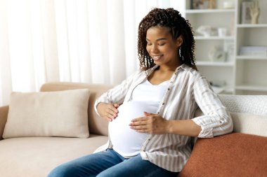 Siyah hamile kadın karnını okşarken gülümsüyor. Evde gebeliğin son evresinde annelik bağı ve sevincinin huzurlu ve samimi anı, kopyalama alanı