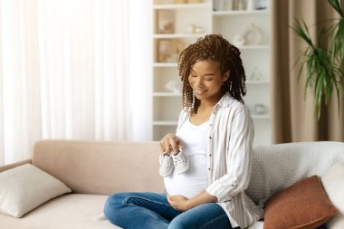 Karnında küçük bebek ayakkabıları taşıyan, yumuşak bir gülümsemesi olan hamile bir zenci kadın. Doğum öncesi yolculukta beklenti, ilgi ve duygusal bağ gösteren sembolik bir an.
