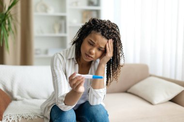 Siyahi bir kadın koltukta oturmuş, endişeli ve hamilelik testi yapıyor. Afrika kökenli Amerikalı bir kadının beklenmedik ya da belirsiz bir şekilde duygusal bir etki yakalaması özel bir ev ortamında sonuçlandı.