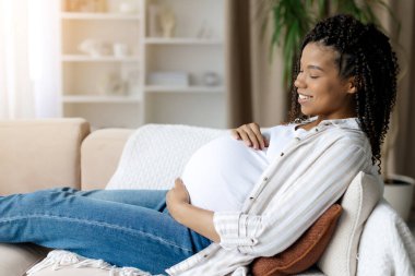 Siyah hamile kadın kanepede dinleniyor karnını tutarken hafifçe gülümsüyor. Huzurlu üçüncü üç aylık dönem anne sevgisini, dinlenmeyi ve duygusal bağlantıyı yansıtıyor.