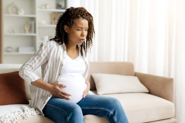 Kasılmalar. Siyah hamile kadın koltukta oturuyor, derin derin nefes alıyor ve karnını tutuyor. Emek hazırlığının gerçekçi tasviri, ağrı yönetimi ve doğum öncesi farkındalık, kopyalama alanı